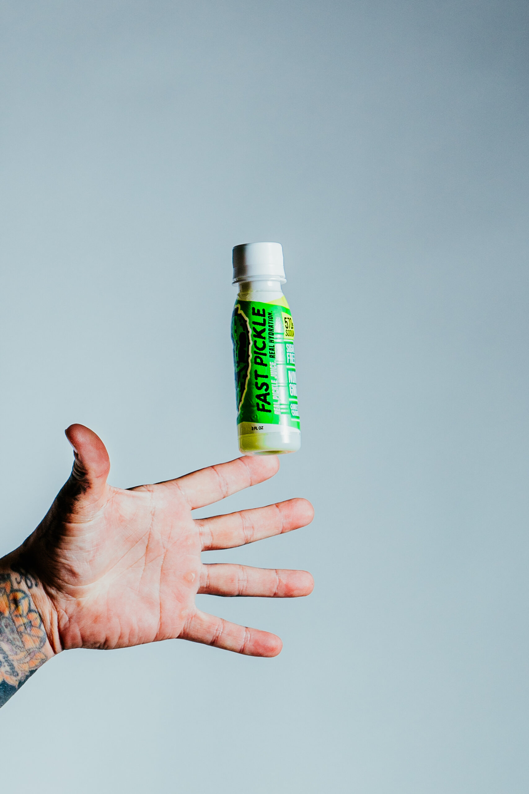 A hand tossing a Fast Pickle 3oz pickle juice shot in the air against a clean grey backdrop — editor pick for the best pickle juice for sports.