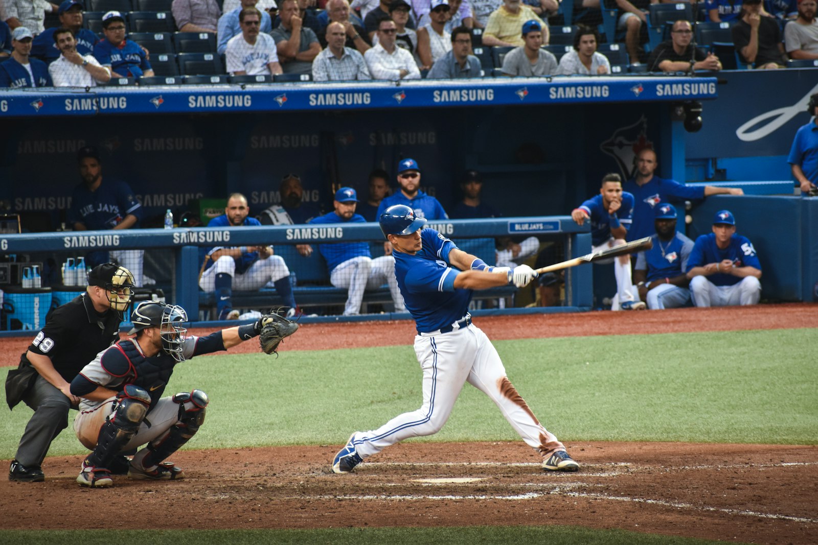 Baseball batter mid-swing during a Major League game with catcher and dugout behind — illustrating the hydration and sodium demands of a long summer baseball season.