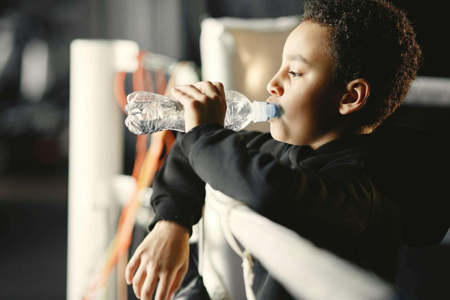 A young boy in a black hoodie drinks water from a plastic bottle in a boxing gym.