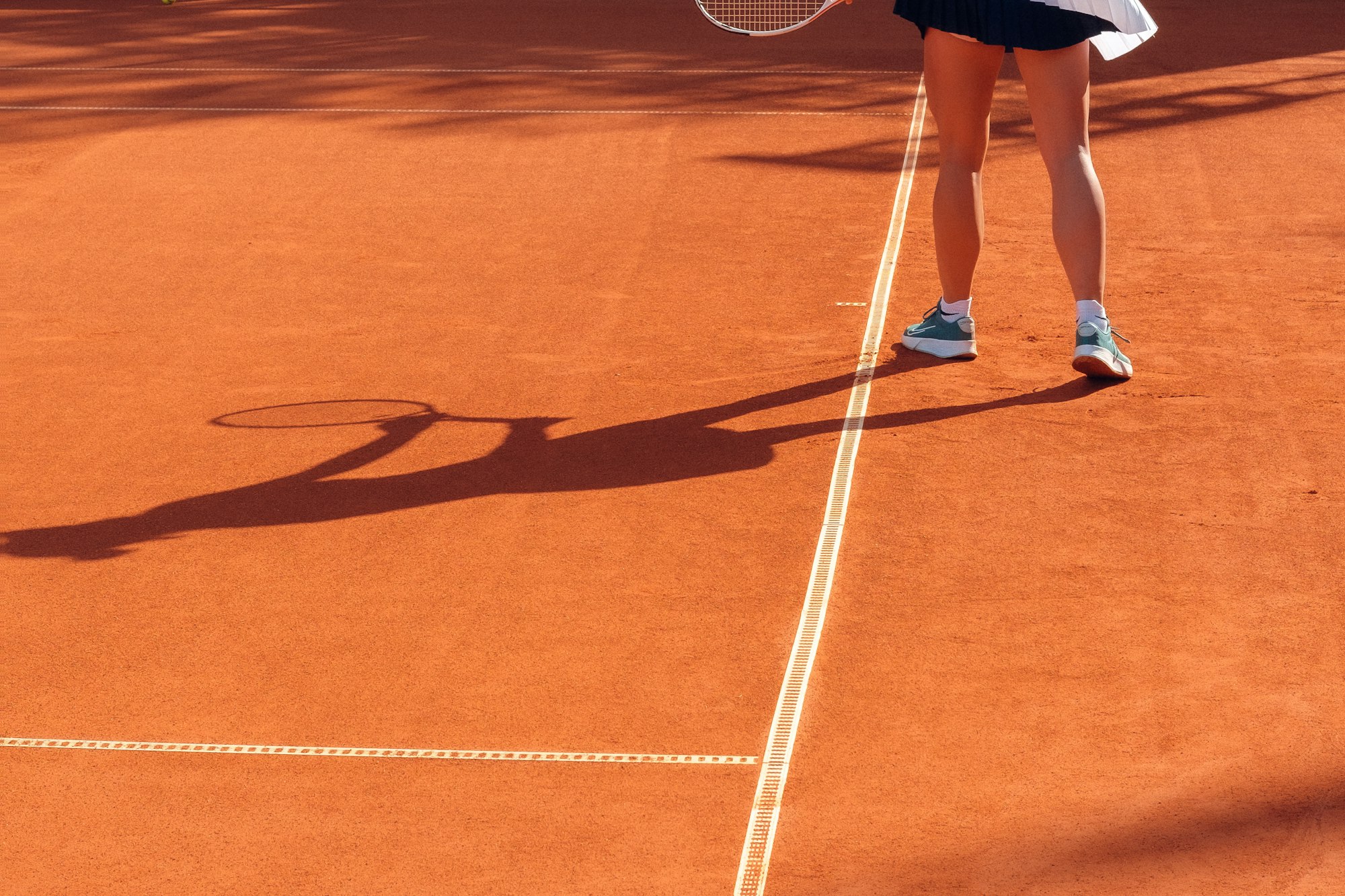 Tennis player standing on a red clay court with a long shadow stretching across the baseline