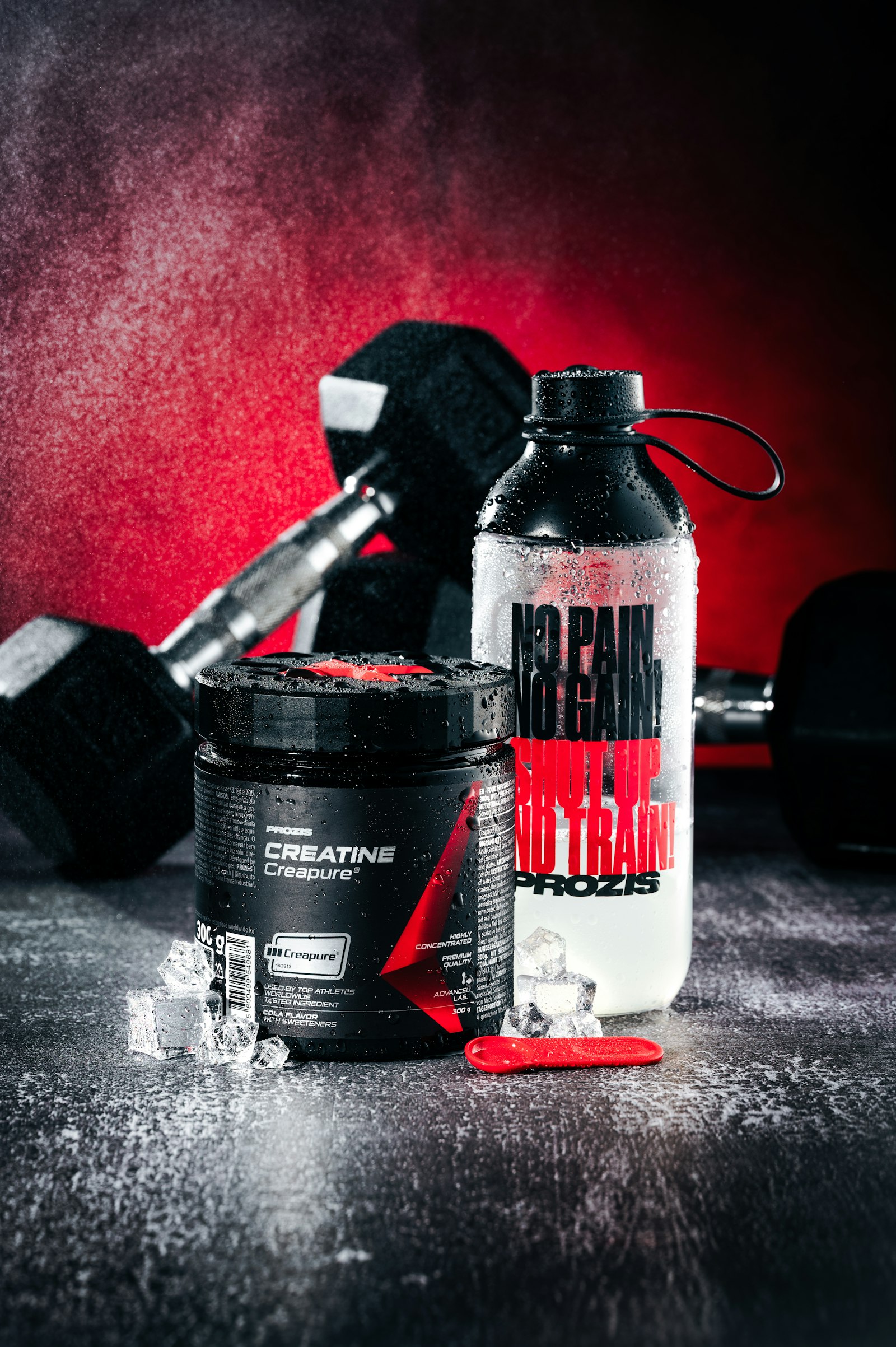 PROZIS Creatine Creapure supplement container and water bottle on dark wet surface with red gym backdrop — illustrating creatine and hydration needs for athletes