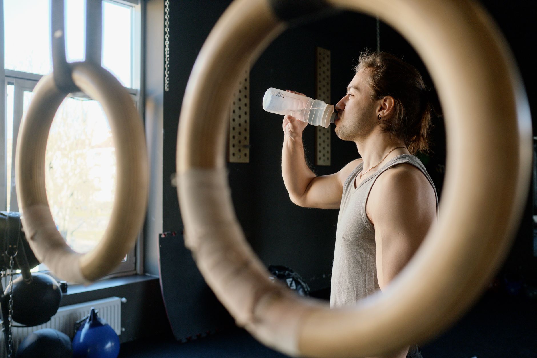 Athletic man drinking water in gym through gymnastic rings for hydration after workout.
