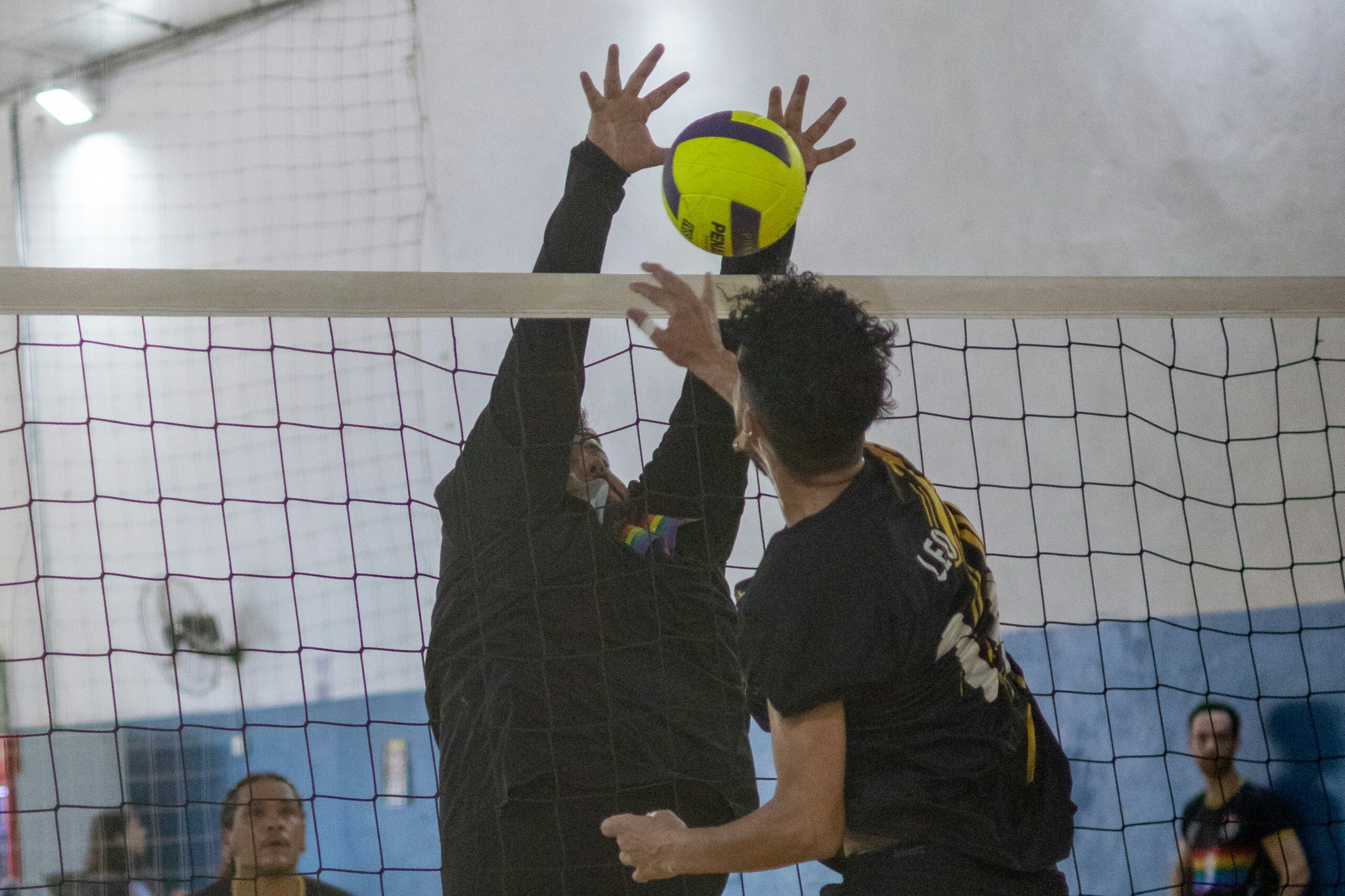 Two men competing at the net during an indoor volleyball match — illustrating the high-intensity sweat demands and hydration needs of volleyball players.