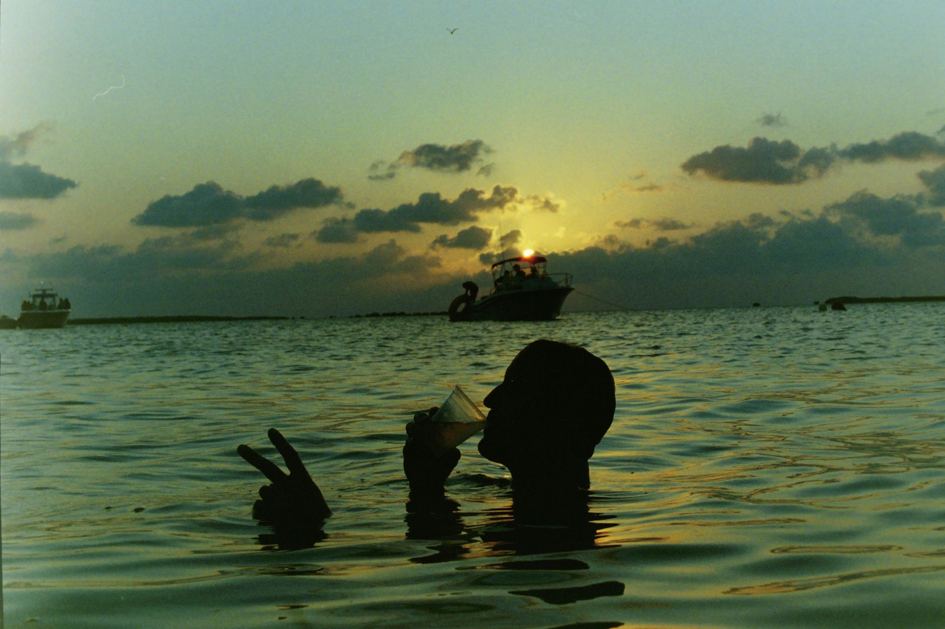 Silhouette of a person enjoying a drink in the sea at sunset, with boats in the background.